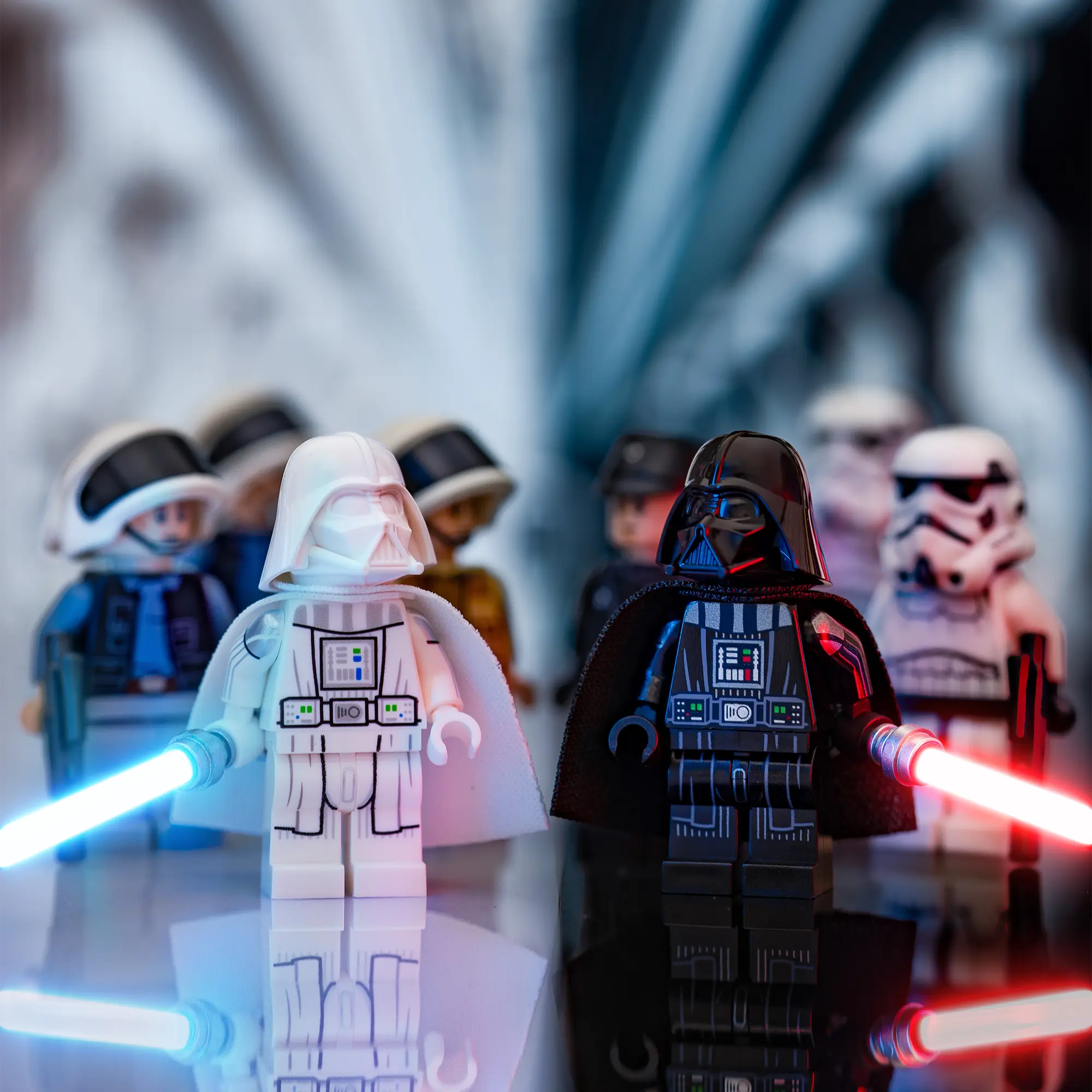 LEGO Darth Vader in black and an alternate white-armored Vader minifigure stand side by side, lightsabers ignited, with Star Wars characters blurred in the background.