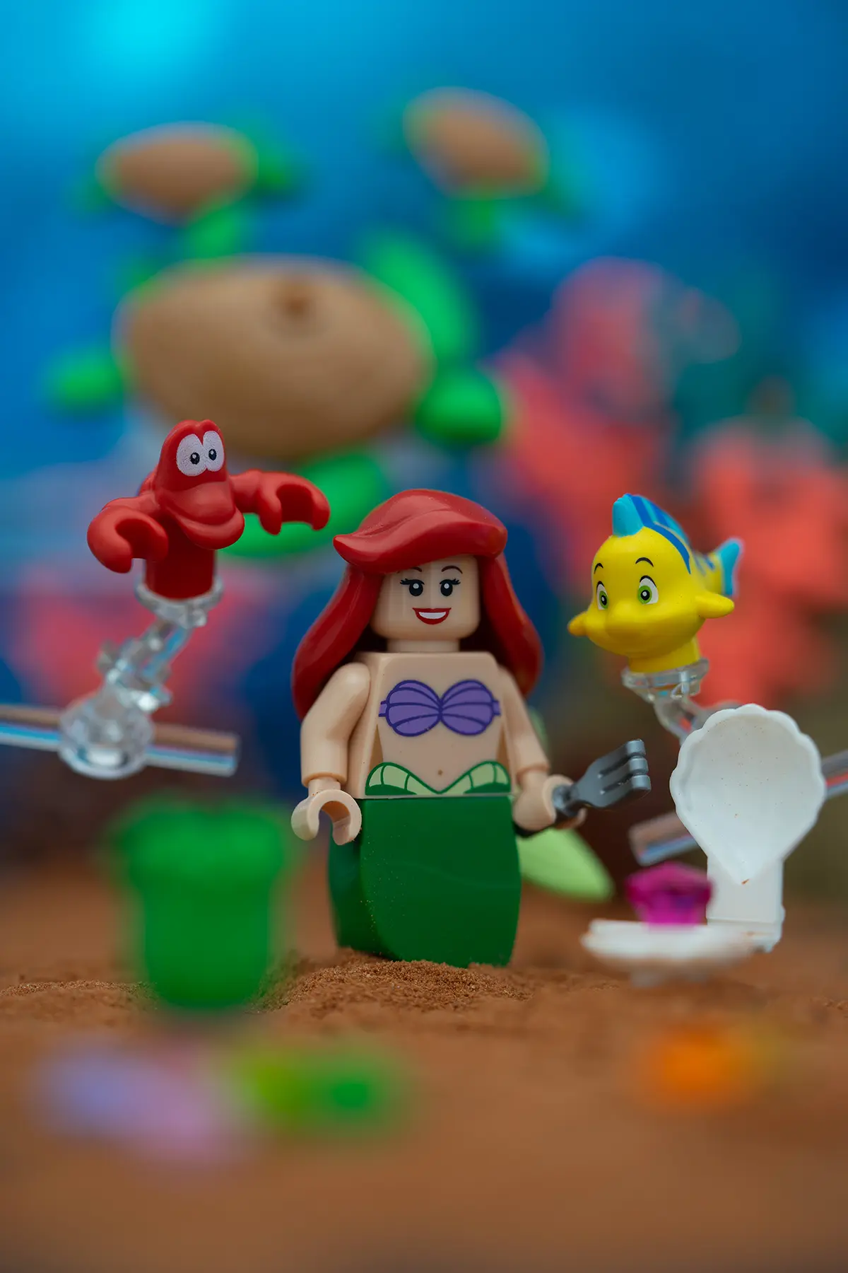 LEGO minifigure of Ariel from The Little Mermaid stands on a sandy ocean floor, flanked by Sebastian and Flounder held up by visible Support bars. The background features colorful corals and sea creatures in soft focus.