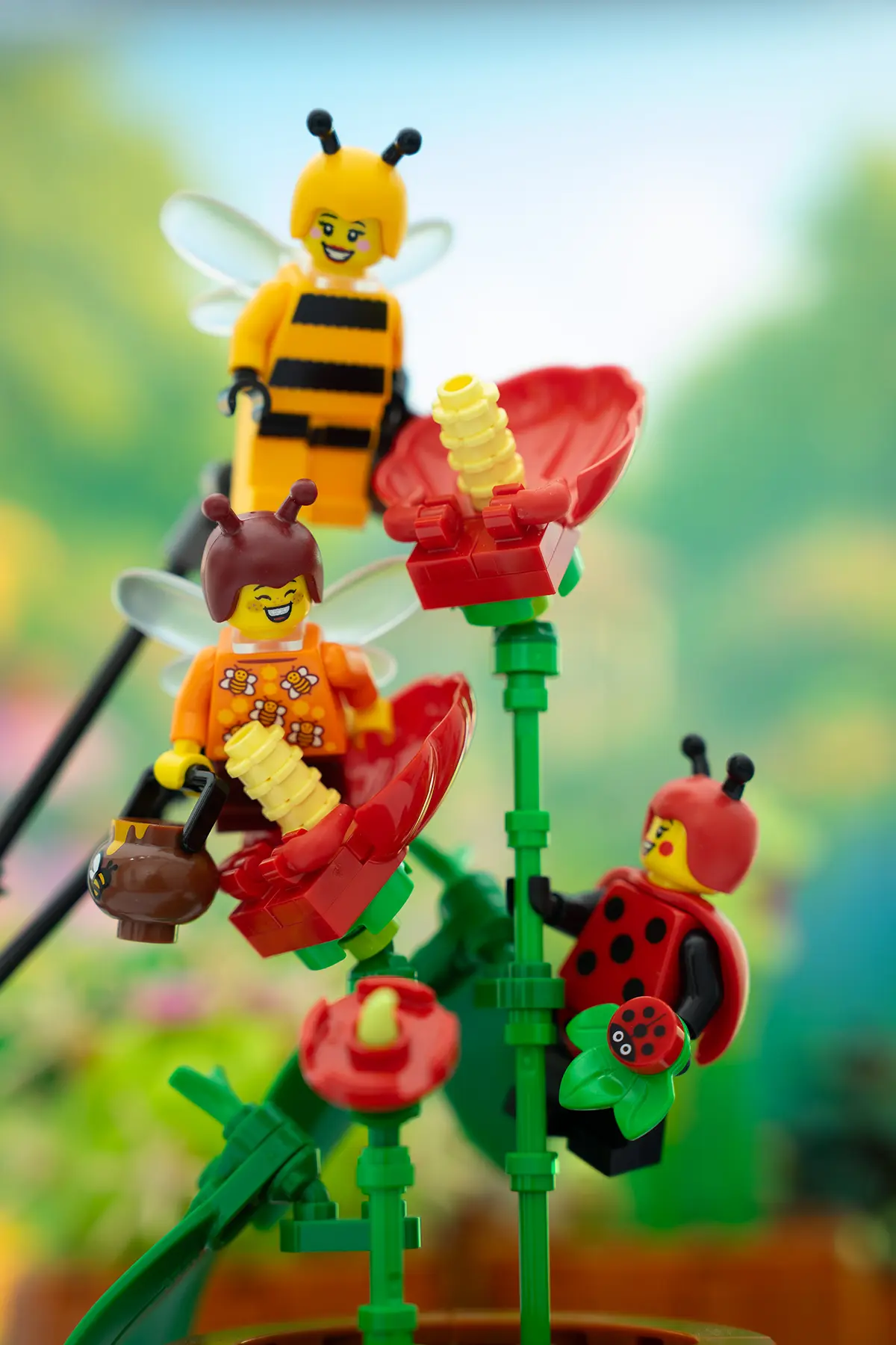 LEGO minifigures dressed as bees and a ladybug pose among red LEGO flowers in a bright, playful garden setup. Clear Support bars and stands are visible, helping position the flying characters