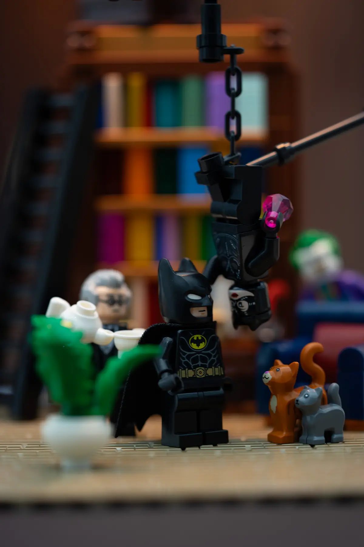 LEGO Batman stands indoors beside a hanging Catwoman minifigure suspended from a black chain, while Alfred serves tea and cats gather nearby. A visible rod supports Catwoman's pose in a warm, book-filled room.