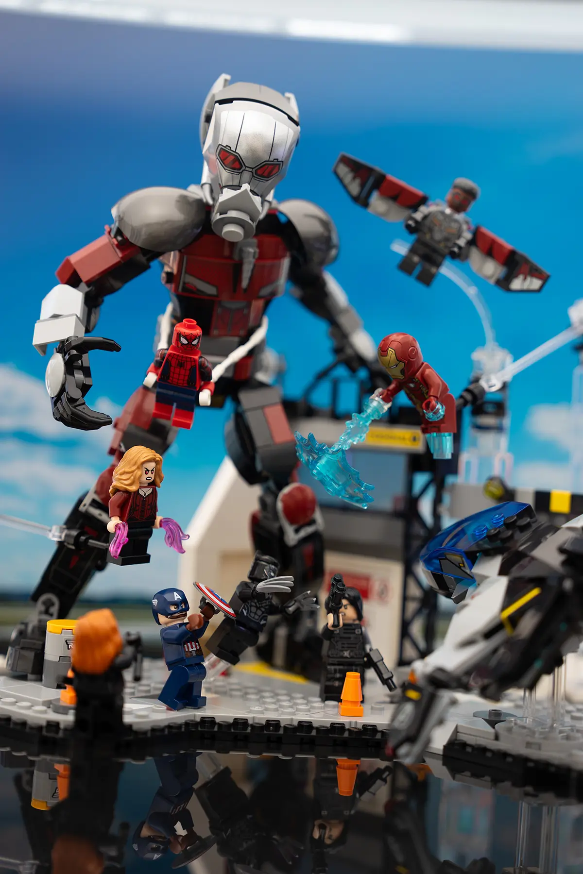 LEGO recreation of the Captain America: Civil War airport battle, featuring Giant-Man, Iron Man, Spider-Man, Falcon, Scarlet Witch, and more. Characters are held up with visible Support bars and stands in a detailed airport diorama under a blue sky.