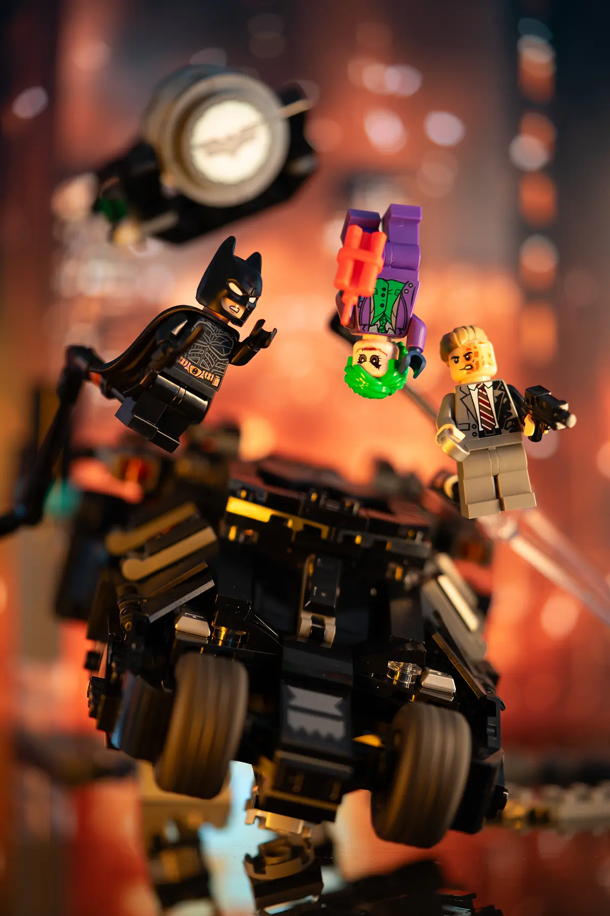 LEGO Batman, Joker, and Harvey Dent minifigures are positioned mid-action above the Batmobile, with Gotham's skyline in the background. The scene includes Support bars for suspension and dramatic lighting.