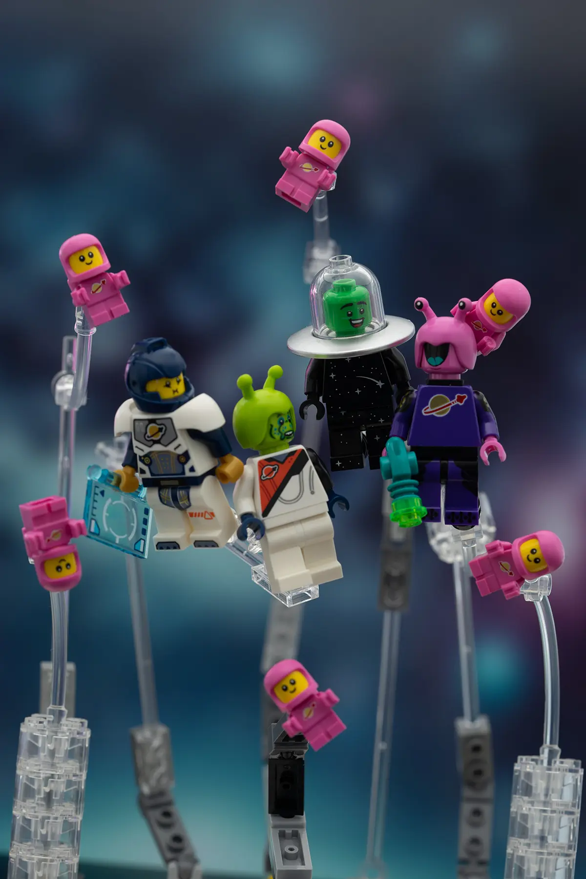 LEGO space-themed minifigures—including astronauts, aliens, and multiple pink baby spacemen—are posed on clear plastic stands in front of a galactic backdrop. The setup elements like rods and clamps are still visible.