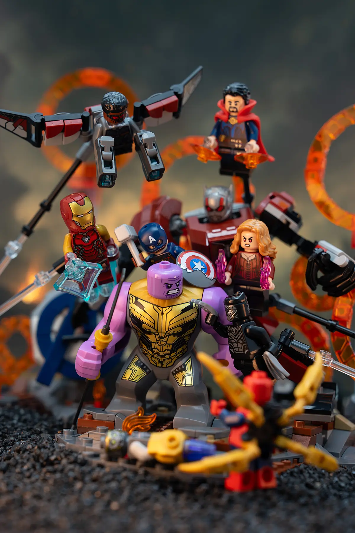 LEGO Avengers minifigures, including Iron Man, Captain America, Black Panther, Scarlet Witch, and others, face off against Thanos on rocky ground. Support rods are visible, and translucent orange portal rings glow in the background.