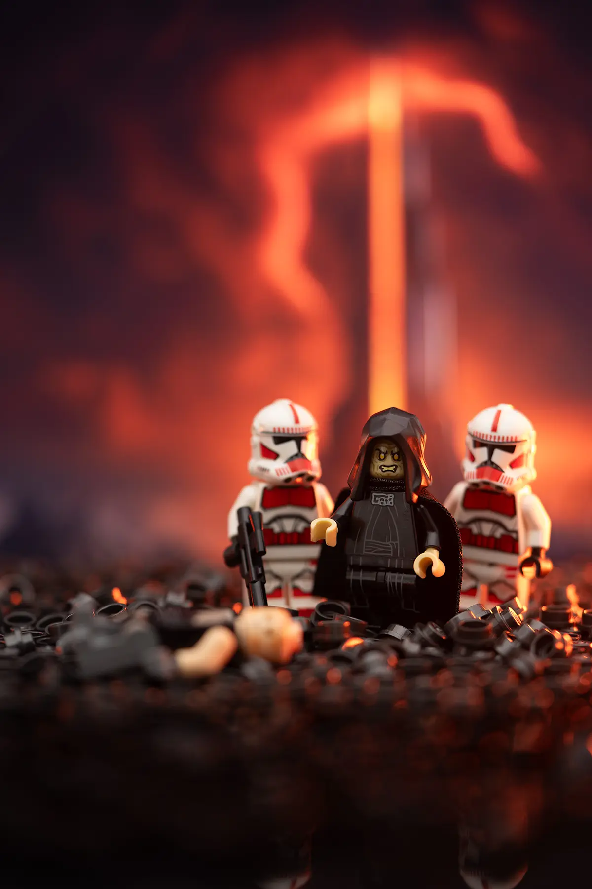 LEGO Emperor Palpatine stands flanked by two Clone Troopers on a scorched, volcanic terrain made of black LEGO pieces. A fallen figure lies at their feet. The background shows a fiery lava flow and swirling smoke, with dramatic lighting and practical effects in place.