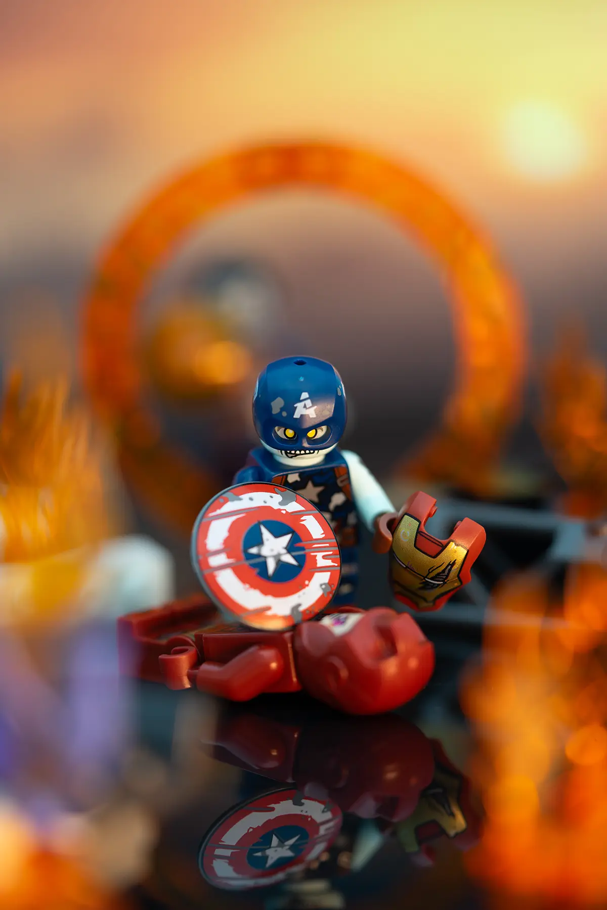 LEGO Zombie Captain America stands over a fallen Iron Man, holding his damaged shield and Iron Man's helmet. The background shows flames and a large glowing ring, with a warm sunset atmosphere and visible support for flame elements.