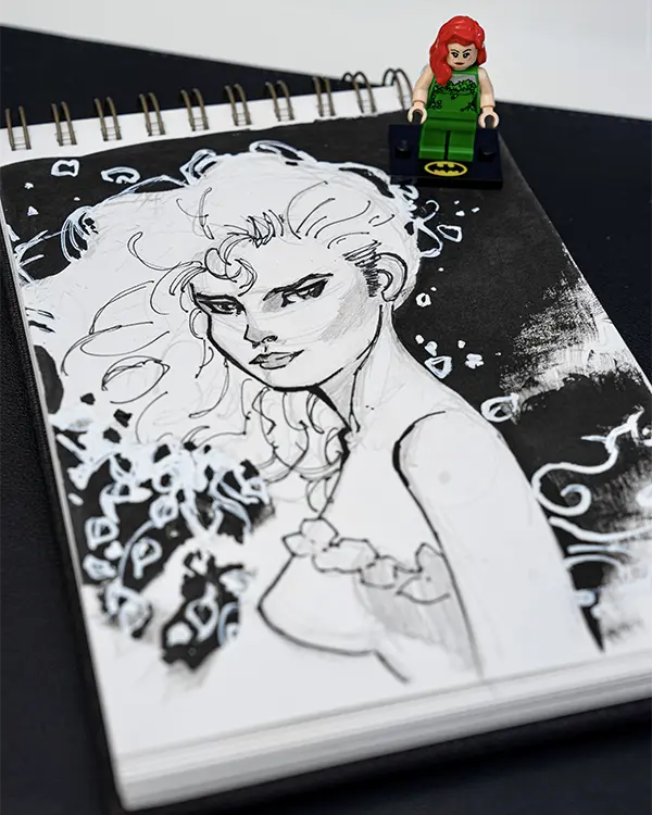 LEGO Poison Ivy minifigure sitting atop a sketchbook featuring a detailed ink drawing of her character.